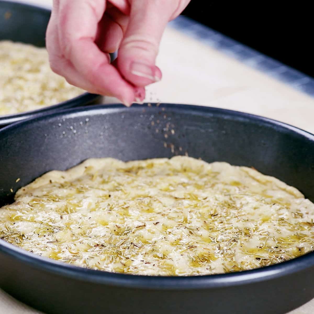 A hand sprinkling black pepper on cottage cheese focaccia dough in a round pan.
