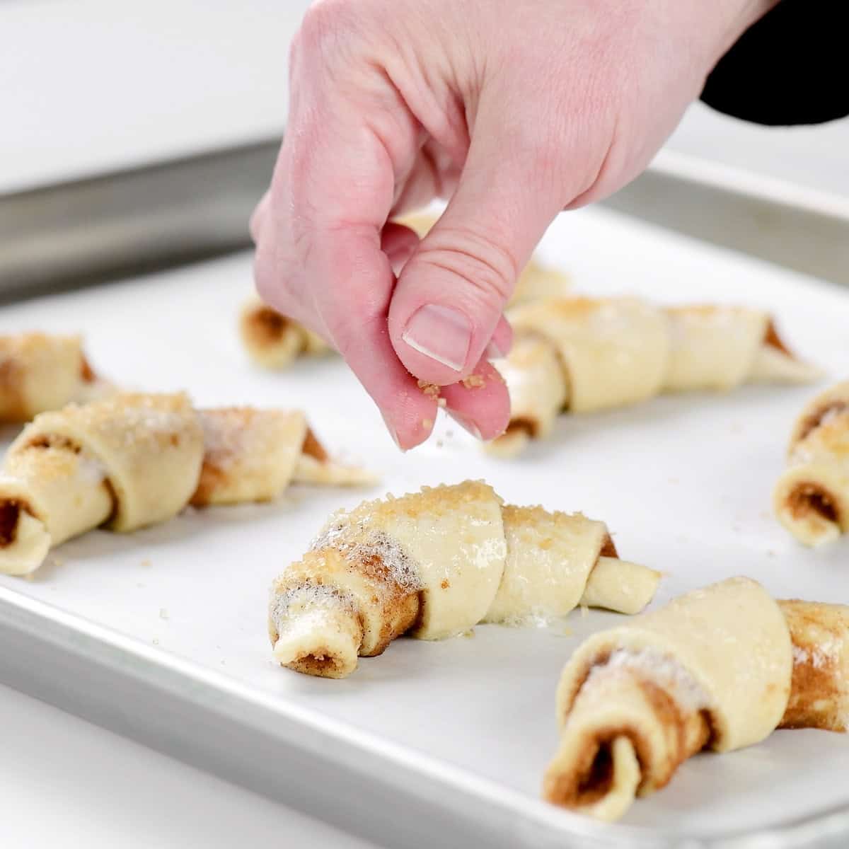 A hand sprinkling coarse sugar on cottage cheese cinnamon crescent rolls.