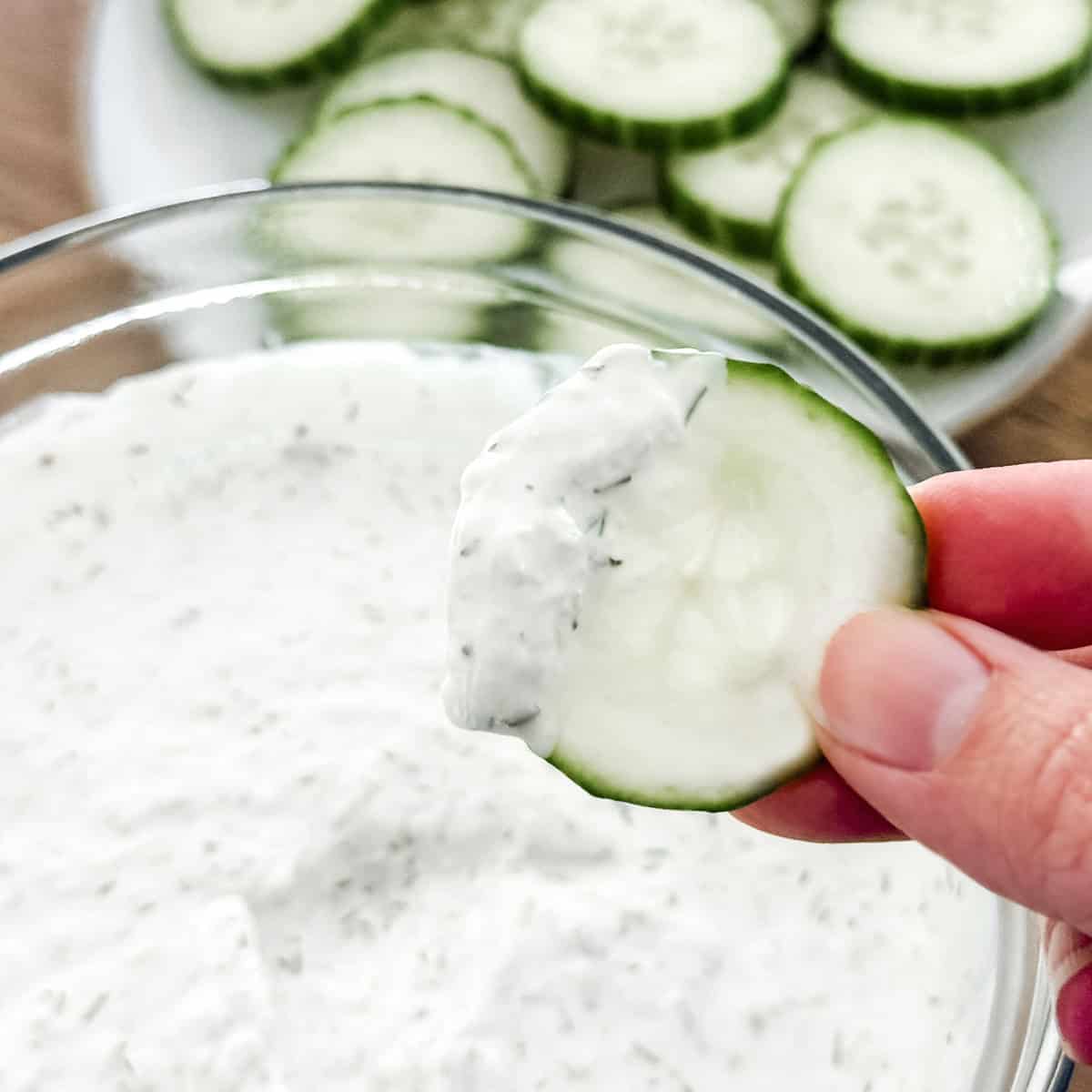 A hand holding a cucumber slice with cucumber dill sauce on it.