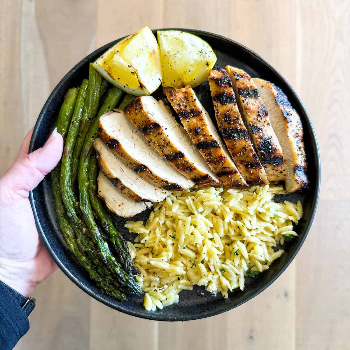 Lemon pepper chicken, orzo, asparagus, and lemon wedges on a black plate.