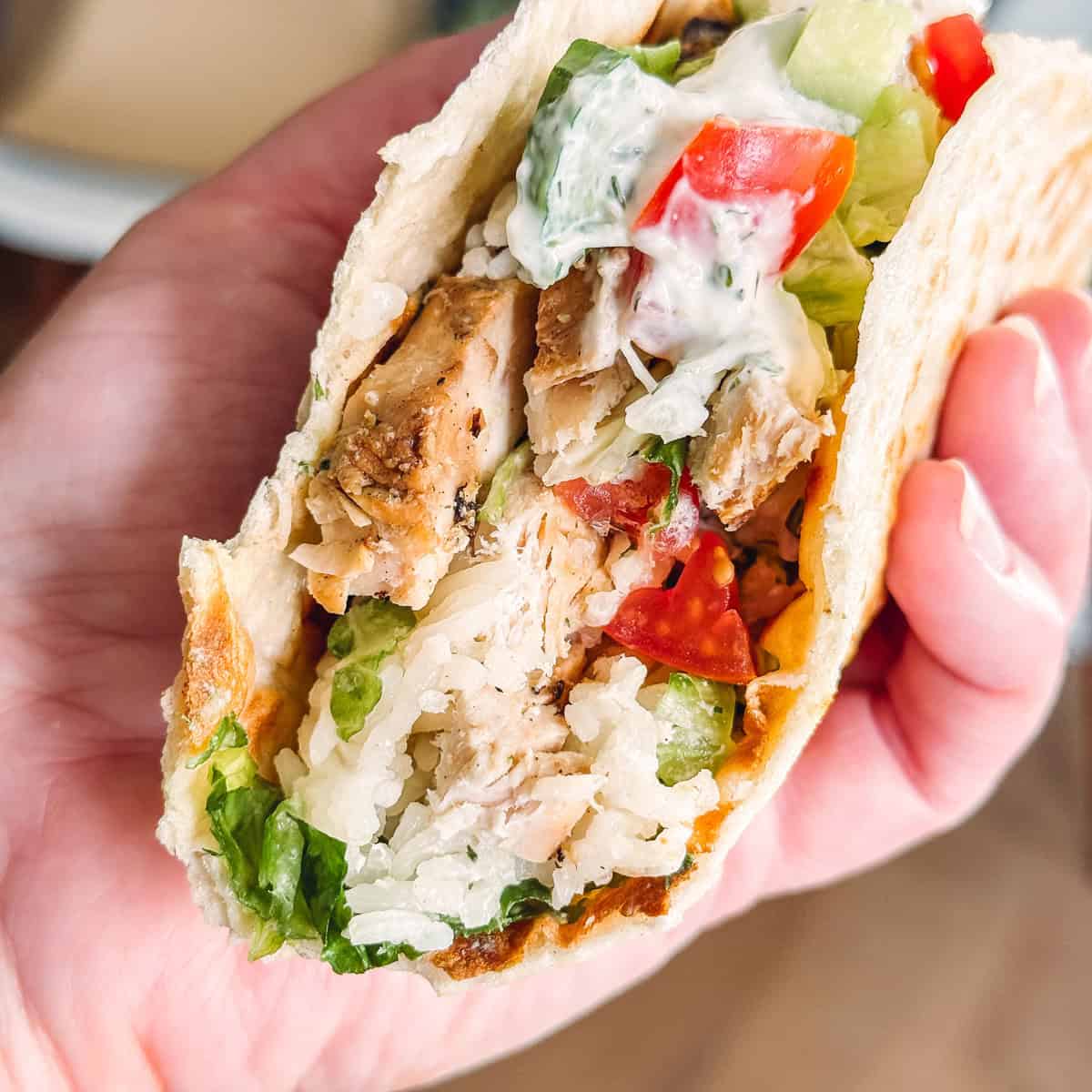 A hand holding a Greek chicken wrap with rice, veggies and sauce.