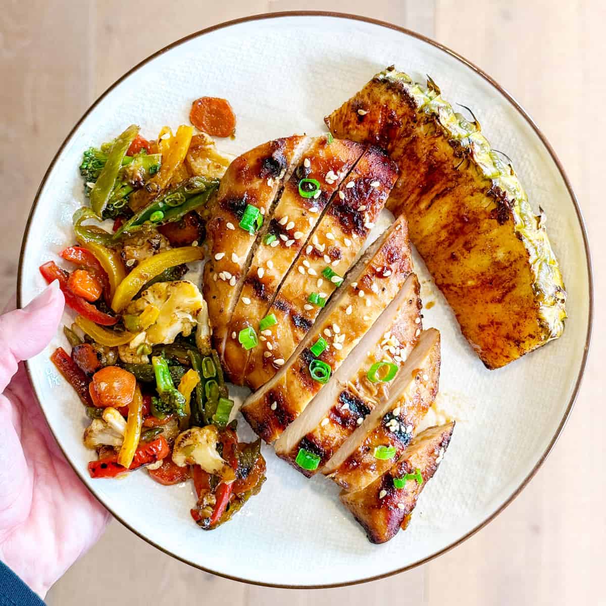 A hand holding a plate with sliced marinated teriyaki chicken, stir fry vegetables, and grilled pineapple.