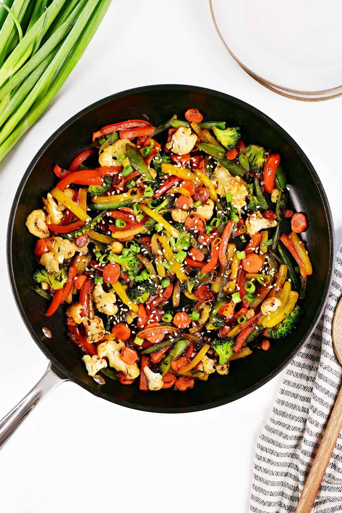 Vegetable stir fry in a skillet, plates, sliced green onions, and a spoon on a dish towel.