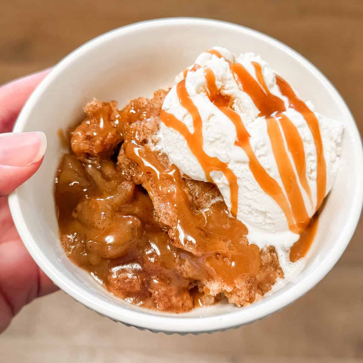 A hand holding a bowl with slow cooker dump cake and vanilla ice cream in it.