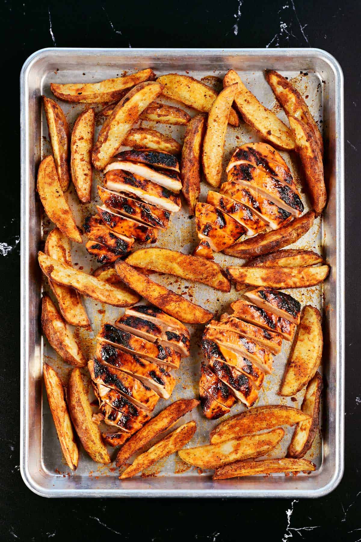 Sheet pan with sliced, marinated buffalo chicken breasts and potato wedges.