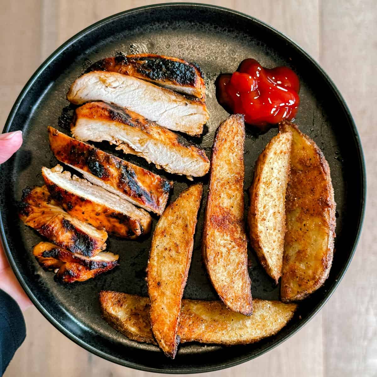 A hand holding a plate with sliced, marinade buffalo chicken and potato wedges.